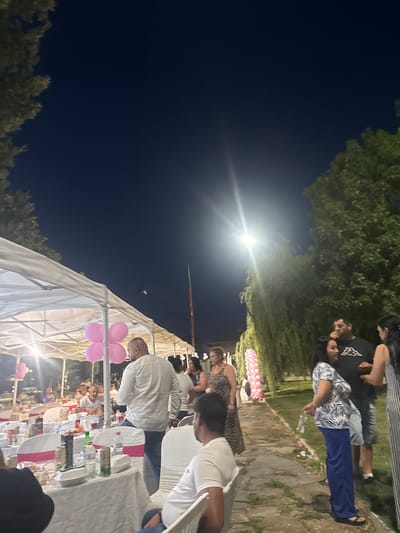 In Golyam Chardak, Bulgaria, an outdoor evening event is underway, possibly a celebration. A white tent covers tables set for a meal with white tablecloths and pink accents. People are gathered around the tables and standing in groups. A man in a white shirt is prominently featured in the foreground, facing away from the camera. Pink balloons are visible, some arranged in a cluster. Trees and foliage surround the area, and the sky is dark. A bright light source, likely a lamp, illuminates the scene. A flagpole is visible in the background.