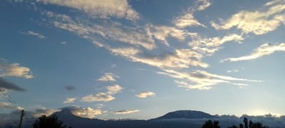 The scene depicts a panoramic landscape captured in Kuku ward, Kenya, at what appears to be either sunrise or sunset. The sky, dominating approximately 70% of the frame, is a clear light blue, accented by numerous scattered cirrocumulus and altocumulus clouds. These clouds exhibit warm, golden-white illumination from the sun, which is outside the frame, suggesting a low angle.
In the mid-ground, two prominent mountains are silhouetted against the lighter horizon. The mountain on the left is a distinct, sharp peak, dark in coloration, with some lighter cloud formations near its base. To the right, a larger, broader mountain displays a flat, wide summit, with visible patches of white, indicative of snow or ice cover. Its lower slopes are obscured by a faint haze or low-lying cloud band.
The immediate foreground includes dark silhouettes of several trees clustered along the horizon line, particularly towards the right. On the far left, a utility pole, dark and slender, extends upwards from the bottom of the frame, carrying multiple overhead wires that traverse diagonally towards the upper left. Two small, dark specks, possibly birds, are visible against the sky; one in the upper right quadrant and another smaller one in the upper left. No people or specific actions are discernible within the frame.