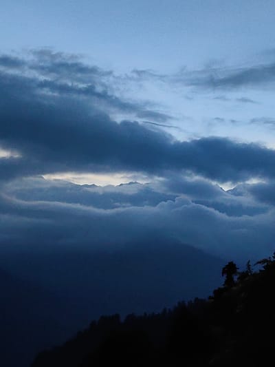 This is a vertical, low-angle shot of a cloudy sky above a dark, silhouetted mountain range, likely taken at dusk or dawn. The sky is a gradient of pale blue at the top, transitioning to darker blues and grays as it meets the clouds. The clouds are dense and layered, with a bright, horizontal band of light breaking through the middle, suggesting the sun is just below the horizon. The mountain range below is a deep, dark silhouette, with the outlines of trees visible along the ridge. The overall impression is one of dramatic natural beauty, evoking a sense of peace and solitude. The scene does not depict any people, specific actions, or urban elements, suggesting a remote, natural setting. The location is stated to be in Siumsa, India, a village in the Lahaul and Spiti district of Himachal Pradesh.