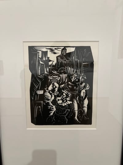 A framed, monochromatic artwork, likely a woodcut or linocut print, is displayed on a white wall. The print, characterized by stark black and white contrast and bold lines, depicts a dynamic urban scene populated by at least five figures. These figures are rendered in elaborate, possibly theatrical or period, attire, featuring ruffled collars, hats, and patterned clothing.
In the foreground, a figure wearing a polka-dotted dress with puffy sleeves kneels or crouches, looking upward. Other central figures stand, one holding a staff-like object, engaged in apparent interactions. A small, dark animal, possibly a dog, is visible near the feet of figures on the right side. The background features a densely packed array of angular buildings, including a prominent structure resembling a church with a steeple on the upper right. Fences, a basket, and other architectural and environmental elements fill the remaining space.
This artwork is on display in a location within Cambridge, United States.