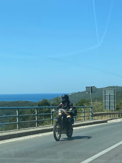 In the city of Seoce, Montenegro, on a clear, sunny day, a person wearing a black helmet and riding a motorcycle is captured from the side in a medium shot. The rider is dressed in a black jacket with yellow accents, black pants, and knee pads. The motorcycle is dark colored with yellow detailing on the front. The motorcycle is on a paved road, and behind the rider, a blue metal railing runs along a concrete barrier. Beyond the railing, lush green trees and vegetation cover rolling hills. In the distance, the blue expanse of the sea is visible. The sky is a vibrant, clear blue with faint white contrails from an aircraft. A sign with a white grid on a dark background is visible to the right of the motorcycle.