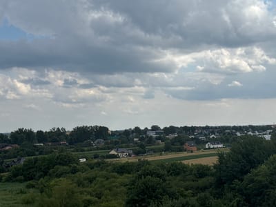 This image depicts a panoramic view of a suburban area, likely on the outskirts of the city of Luboń, Poland, under a cloudy sky. The landscape is characterized by rolling green hills covered in dense trees and interspersed with agricultural fields. Several residential houses are visible, varying in architectural style and color, with some featuring red-tiled roofs and others white or grey facades.
In the foreground, a thick expanse of dark green trees dominates the lower portion of the image, suggesting a natural or park-like setting. Beyond this, the terrain rises gently, revealing a patchwork of fields, some appearing to be cultivated with crops, possibly wheat or barley, in shades of yellow and brown. Scattered among the greenery are numerous houses, some clustered together to form a small village or neighborhood, while others stand more isolated.
One notable building in the mid-ground is a light-colored house with a dark roof, situated on a paved area with a few cars parked outside. Another house, partially constructed from red brick, is visible to the right of this. Further into the distance, more houses and buildings become visible, hinting at a larger urban or semi-urban area beyond the immediate landscape. A white van or truck is parked amongst the trees on the left side of the mid-ground.
The sky is filled with a dramatic display of clouds, ranging from dark, heavy masses to lighter, scattered formations. The overall lighting suggests it may be late afternoon or a day with overcast conditions. There are no visible people in the image. The scene conveys a sense of rural tranquility combined with the presence of residential development. The overall impression is of a typical suburban or semi-rural environment in a temperate climate.
