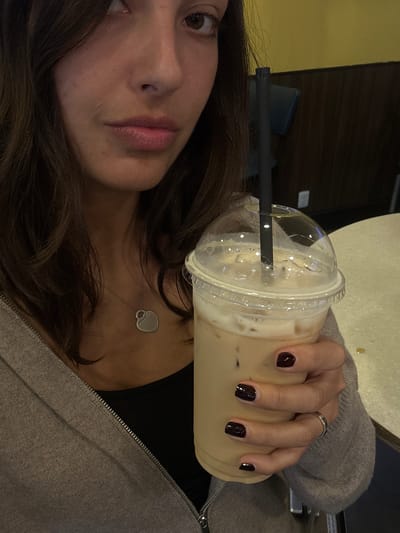 An overhead, slightly angled selfie captures a young woman indoors, likely in Veracruz City, Mexico, holding an iced beverage. Her face, with dark brown hair, is visible on the left side of the frame, looking towards the camera with a neutral expression. She is wearing a grey zippered sweater or hoodie, and a silver heart-shaped pendant necklace rests on her chest. Her right hand, adorned with a silver ring on the ring finger and dark, possibly burgundy, nail polish, firmly holds a clear plastic cup. The cup contains a creamy, light brown iced drink, presumably coffee or a similar beverage, and has a domed lid with a black straw inserted. The background is blurred, showing glimpses of yellowish and dark brown surfaces, suggesting an indoor setting like a cafe or casual eatery. There is no visible text in the image.