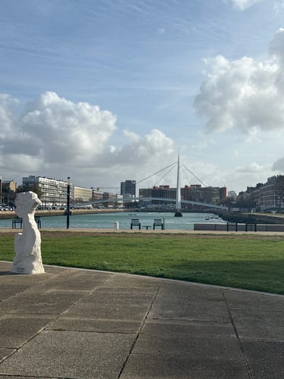 The image depicts a daytime urban waterfront scene in Le Havre, France, looking generally east across the Bassin Vauban. In the foreground, a grey concrete paved path transitions into a manicured green lawn. Positioned on the pavement to the left is a white, abstract stone sculpture, appearing to be a stylized human head or bust, facing towards the right.
Beyond the lawn lies a body of turquoise-colored water, likely a basin or canal. Two dark-colored benches with backs are situated on the water's edge, facing the water. A white bollard is also visible near the benches. Several small boats are moored on the opposite side of the water.
Dominating the middle ground is the Pont Patrick Devedjian (Pont de la Porte Océane), a distinctive white cable-stayed bridge with a single central pylon, spanning the water. The bridge's deck is supported by multiple cables radiating from the pylon.
The background consists of the city of Le Havre. Multi-story buildings, some residential with balconies and others commercial, line the distant shore on both sides of the basin and beyond the bridge. Vehicles are visible on roads adjacent to these structures. The sky overhead is partly cloudy, with scattered white cumulus clouds against a light blue backdrop. No individuals are present in the visible area.