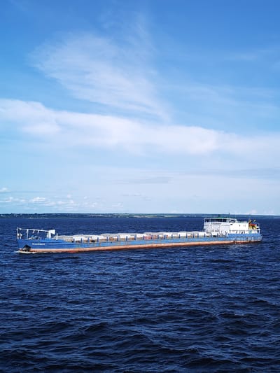 A cargo barge, identified by the name "ВЛАДИМИР" on its bow, sails on a dark blue, choppy body of water, likely a river or lake. The barge is predominantly blue with a reddish-brown waterline. It is loaded with white rectangular containers stacked along its deck. The barge is positioned in the middle ground of the image, moving from left to right. In the background, a distant, green shoreline with scattered buildings and trees is visible under a bright blue sky with scattered white clouds. The environment suggests a daytime scene under a clear sky. The location is the city of Myaksa, Russia.