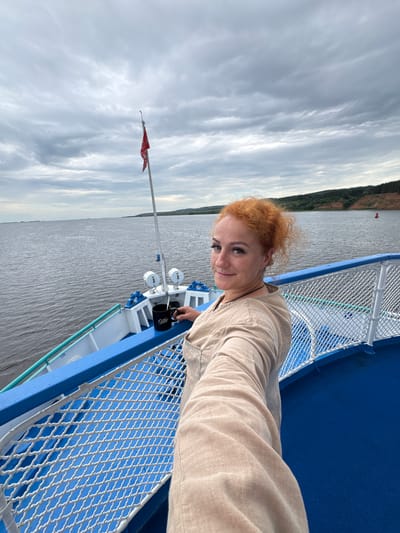 A woman with curly red hair is taking a selfie on the deck of a boat. She is wearing a light brown shirt. The boat has a blue railing and a white net fence. In the background, there is a wide body of water, a cloudy sky, and a tree-covered shoreline with a red buoy visible in the distance. The image appears to be taken in a natural setting, likely on a river or large lake. It is not possible to determine the specific location within the city of Menzelinsk, Russia, from the visual information provided.