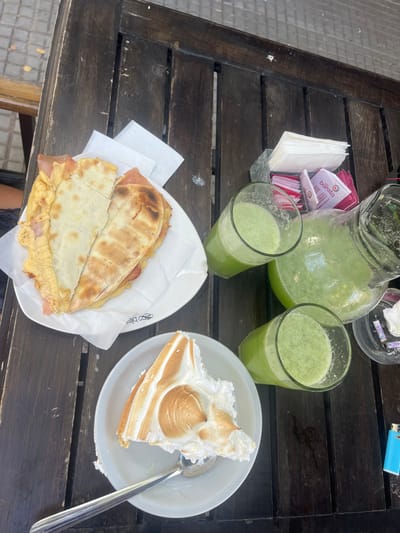 This image captures a tabletop scene at an outdoor establishment in City Bell, Argentina. A dark brown wooden slatted table is laden with food and beverages. On the left, a white oval plate displays a folded flatbread, possibly *fainá* or *fugazzeta*, filled with ham and a yellow ingredient, resting on a crumpled white paper napkin. Below this, a white circular plate holds a partially consumed slice of dessert, notable for its tall, toasted meringue topping, with a silver spoon resting alongside it. To the right, a clear glass pitcher is approximately two-thirds full of a light green, frothy beverage, accompanied by two individual clear glasses, each containing a similar quantity of the same drink. In the upper right, a metal napkin dispenser contains white paper napkins and several pink "barista" branded sugar packets. Adjacent to this, a clear glass ashtray holds several cigarette butts and a used tissue. A light blue Bic lighter is positioned on the table next to the ashtray. No individuals are visible in the frame.