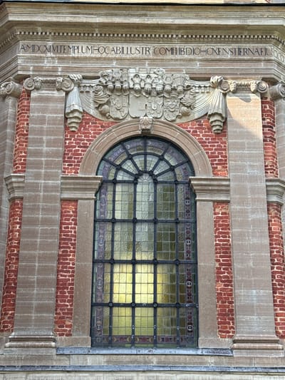 The image shows a close-up, straight-on, eye-level exterior architectural detail of a building in Askersund, Sweden. The structure features red brick walls with light-colored stone pilasters and decorative elements. Above a large, arched stained-glass window with a dark metal frame, there is a stone frieze with carved inscription. To the left and right of the frieze are decorative stone carvings. The window is divided into many small panes, with the glass appearing to be a yellow or golden hue. The inscription on the frieze is in Latin and appears to be a dedication or historical marker. The overall impression is of an old, possibly historical building with ornate architectural features.