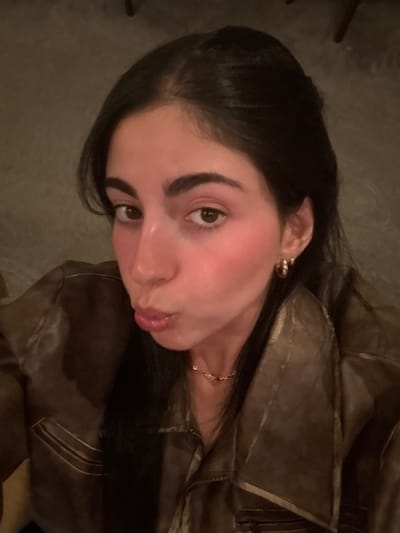 A female subject is depicted in a close-up, chest-up shot. She has dark hair, dark brown eyes, and prominent eyebrows. Her skin appears fair with a pinkish blush on her cheeks. Her lips are pursed in a "kissing" expression, and she looks directly at the camera. She is wearing a dark brown or olive-green jacket, possibly leather or faux leather, featuring a wide collar and horizontal seam details on the chest. Accessories include small gold-colored hoop earrings in her visible right ear and a delicate gold-colored chain necklace. The background is uniformly dark and out of focus, offering no discernible features or objects. Lighting is dim and warm, consistent with an indoor evening setting. The image provides no visual information to identify the location as Rum Cay, Bahamas, or any specific city.