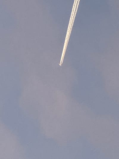 The image shows a clear blue sky with faint clouds. A jet airplane is visible, leaving a prominent contrail that stretches diagonally across the sky. The airplane itself is a small, distant speck against the vastness of the sky. The scene appears to be an aerial view, possibly taken from another aircraft or a high vantage point. There are no discernible urban or natural landmarks visible in the image, making it impossible to determine a specific location within Cuxton, United Kingdom, or any other city.
