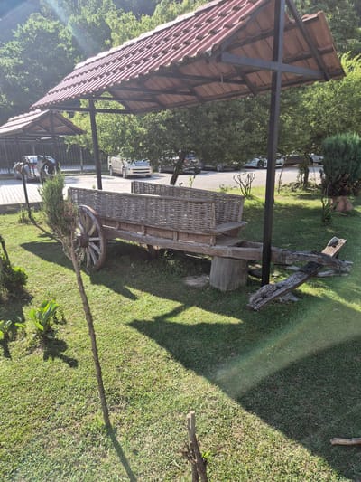 The image depicts an outdoor scene in Bzypta, Abkhazia, bathed in daylight, likely morning or afternoon given the bright sunlight casting distinct shadows. The setting appears to be a park or garden area, characterized by lush green grass and mature trees providing a natural, shaded environment.

The focal point of the image is an antique wooden cart with woven sides, resting on a large wooden wheel. This cart is positioned on the grass, next to a weathered wooden structure that resembles part of an old mill or some other rustic machinery. The cart and its accompanying wooden element suggest a historical or decorative display within the location.

In the background, a modern carport with a red tiled roof is visible, supported by dark metal beams. Beneath the carport, several cars are parked, indicating a public or commercial space. The contrast between the old cart and modern vehicles highlights a blend of old and new within the landscape.

Beyond the carport, dense green foliage from trees fills the upper portion of the frame, suggesting a verdant and perhaps somewhat secluded location. The overall impression is one of a peaceful, well-maintained outdoor area, possibly a tourist attraction or a pleasant resting spot. There is no visible text in the image.