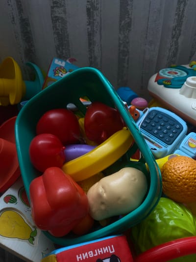 In an indoor setting, likely a child's play area, a collection of plastic toy food items is piled together. The dominant colors are bright and varied, with a prominent teal basket holding several red toy vegetables, including what appear to be tomatoes and a bell pepper. A yellow banana-shaped toy is also visible, along with a light-colored toy potato. Other toys scattered around include a blue toy calculator, a toy orange, and a green toy cabbage. The background features a textured, light-colored curtain with vertical stripes and a hint of a patterned tablecloth or playmat with fruit designs. The scene is well-lit, with shadows suggesting a light source from the right side of the frame. The toys appear to be for a young child's play and are not associated with any specific activity or interaction. The location of Пешнево, Russia is not determinable from the visual information present in the image.