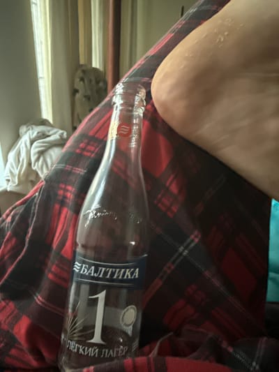 In an indoor setting, a clear glass bottle of Baltika No. 1 Lager is placed on a red and black plaid blanket. The bottle is positioned in the foreground, with a person's foot and lower leg, clad in the same plaid blanket, occupying the right side of the frame. The background is blurred but suggests a domestic environment with curtains and a draped cloth. The scene is observed from a close-up, angled perspective, focusing on the bottle and the blanket. There is no information to indicate this location is in Lesnoi Gorodok, Russia.