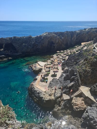 The image depicts a coastal scene near Plakias, Greece. A rocky cliff descends towards the turquoise waters of the Mediterranean Sea. At the base of the cliff, a small, partially constructed beach area is visible, furnished with several straw umbrellas and lounge chairs. Numerous individuals populate the beach area, some sunbathing under the umbrellas, others walking along the concrete pathway. Several people are swimming and snorkeling in the clear, turquoise water. A set of stairs carved into the rock leads down towards the beach area. The horizon reveals a clear blue sky meeting the expansive ocean. The overall environment suggests a summer day at a coastal resort or public beach area. The image is oriented vertically with some minor tilting.