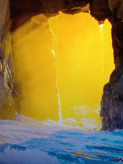 The image displays a natural rock formation or cave entrance situated in the city of Olambe, Nigeria. A large volume of intense, bright yellow light emanates from an external opening, illuminating the central area. Dark, rugged rock walls form an archway at the top and frame the scene on the left and right. Visible shafts of light penetrate through what appears to be mist or spray within the opening. At the base of the rock formation, a body of blue water with significant white foam and crests indicates active wave or current movement. Water droplets or a thin stream are observed descending along the left rock face. No individuals or man-made objects are present within the frame.