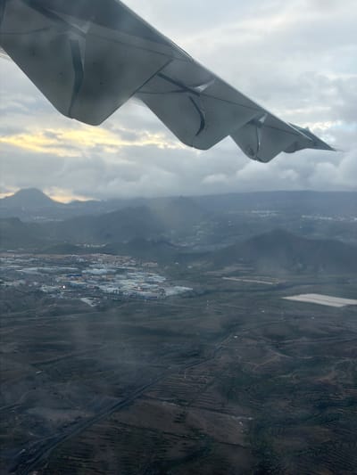 The image presents an aerial view captured from the window of an airplane, looking down at a landscape with a prominent aircraft wing in the foreground. The perspective is rotated 90 degrees counter-clockwise from a typical horizontal view, showing the wing on the left and the ground stretching to the right.
The left side of the frame is dominated by the light gray wing of the aircraft, featuring a distinct wingtip device (likely a sharklet or winglet) pointing upwards and slightly forward. Sections of the wing's trailing edge, possibly flaps or slats, are visible, suggesting the aircraft may be in a phase of flight like ascent or descent rather than cruise. A small, indistinct logo or text, possibly resembling a stylized "W" or "M," can be observed on the visible underside of the wingtip device.
Below and to the right of the wing, the diverse landscape of Granadilla de Abona, Spain, unfolds. In the midground, a significant urbanized or industrial area is visible, characterized by numerous light-colored, predominantly rectangular roofs, likely warehouses, factories, or commercial buildings. These structures, along with some darker roads or ground, suggest a modern development, potentially an industrial estate or port facility consistent with the Port of Granadilla. Some artificial lighting appears to be active within this complex, casting reflections or providing illumination.
Beyond this developed area, the terrain transitions into a more natural, arid, and hilly or mountainous landscape. Faint lines and patterns suggest roads, tracks, or terraced agricultural plots winding through the dark, rough ground. The overall weather appears partly cloudy or overcast, with patches of brighter, yellowish light breaking through the clouds on the horizon, hinting at either early morning or late afternoon. The scene conveys a sense of observation from above, capturing the interplay between natural landforms and human development under diffused light.