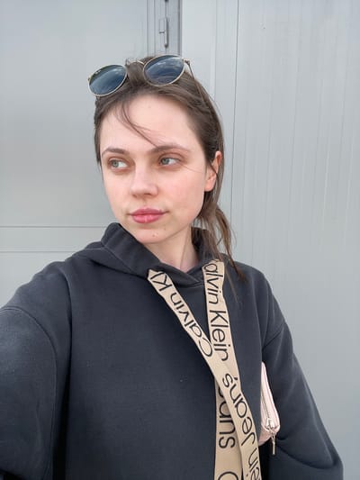 A woman poses for a selfie. She is wearing round sunglasses in her hair, a black hoodie with a Calvin Klein branded strap. She is in front of a light gray wall. The location is undetermined.