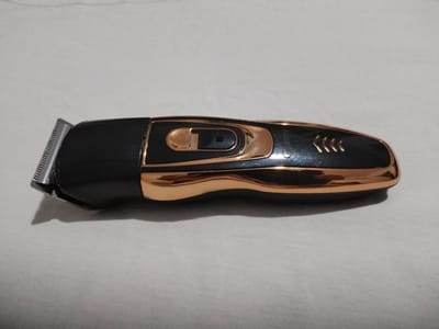 This is a studio photograph of a black and gold electric hair clipper, likely a cordless model, lying on a textured, off-white surface that appears to be fabric. The clipper is positioned horizontally, with its metallic cutting head on the left side of the frame. The body of the clipper is predominantly black, with a glossy, metallic gold trim that curves along its length. This gold trim forms an ergonomic grip area and has a series of three small, raised chevron-like markings towards the right end. In the center of the black body, there is a prominent oval-shaped button or switch, also outlined in gold, with a darker insert, possibly indicating its function. The brand name "GREEEMY" is visible in small, white lettering on the gold trim towards the right end of the clipper. The lighting suggests it's an indoor setting, with a soft shadow cast by the clipper on the surface, indicating the light source is slightly above and to the left. There are no people present in the image, and the focus is solely on the grooming device. The overall impression is a clean, product-focused presentation.