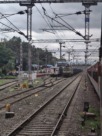 The image shows a railway yard with multiple tracks under a cloudy sky. In the foreground, several sets of train tracks run parallel, covered with gravel and debris. A red and grey train carriage is visible on the right side of the frame. In the middle ground, a green and yellow locomotive is positioned on one of the tracks, pulling a long line of blue cargo wagons. To the left of the locomotive, another train, appearing white and grey, is stationary. A signal pole with an 'X' symbol is visible on the left side of the image. In the background, trees and railway station buildings with blue roofs can be seen. The scene is consistent with the railway station in Khandwa, India, a significant railway junction. The image captures a typical day at the railway yard, with trains parked and ready for operation.