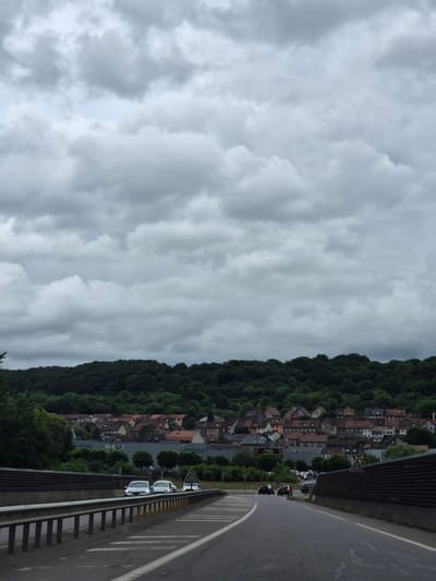 The image displays an elevated view of Audun-le-Tiche, France, under a heavily overcast sky. The scene is dominated by a sweeping grey sky filled with thick, textured clouds, suggesting a cloudy day with muted light.
In the mid-ground, a dense, dark green forest covers a hillside, stretching across the frame horizontally. At the base of this forested slope, a residential area unfolds, characterized by numerous houses with distinct reddish-brown tiled roofs. Streets and parking areas are visible among the buildings, with several cars parked within the town. On one building, the partially obscured text "shop's" is discernible, and another appears to have "20C" or "200" on its facade.
The foreground features a wide, curved asphalt road, likely a bridge or elevated route, extending from the bottom right towards the center. It is bordered by a metal guardrail along its outer edge. Road markings, including solid white, dashed white, and solid yellow lines, are clearly visible. A few vehicles are present on this road: a white car is parked partially off the road near the guardrail, another light-colored car is further ahead, and a dark car is driving away from the viewer. Road signs, including a triangular warning sign indicating a sharp curve and a blue circular information sign, are also visible alongside the road.
The overall impression is one of a quiet, somewhat subdued daytime scene due to the overcast weather.