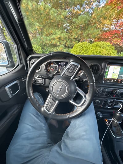 The image displays a first-person perspective from the driver's seat of a Jeep vehicle, stationary in Wellesley, United States. A person's legs, clad in light blue athletic pants, are visible beneath the black leather steering wheel, which features brown stitching and a central "Jeep" emblem. The dashboard ahead includes a digital instrument cluster and a central infotainment screen displaying a map interface. To the right, a gear shifter is visible, with a white charging cable connected. Through the windshield, a bright outdoor scene shows mature trees with green, yellow, and red autumn foliage, along with dense green bushes, indicating a clear, sunny daytime environment during the fall season.