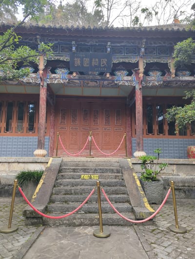 The image depicts the entrance of the Erhai Gate, a historic structure located in Dali City, China. A set of stone stairs leads up to a traditional Chinese building with ornate wooden doors and intricately decorated eaves. Red velvet ropes and golden stanchions are positioned in front of the stairs, suggesting a controlled access or a preserved area. The building's architecture features carved panels, colorful paintings, and wooden pillars. The surrounding environment includes trees and foliage, indicating an outdoor setting. The overall scene conveys a sense of history and cultural significance.