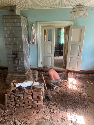 A man is kneeling on the floor of a room undergoing renovation in Бучацінскі сельскі Савет, Belarus. He is shirtless and wearing grey sweatpants and blue sandals. The floor is covered in dirt, debris, and broken bricks. A pile of red bricks is stacked to his left, with a shovel and white foam sheets on top. To his right, another shovel leans against the wall.
The room has pale blue walls and a white paneled ceiling. A large, grey tiled stove with decorative metal inserts stands prominently on the left side of the image, built on a brick base. A pink patterned garment hangs on a hook to the right of the stove. In the background, a set of double doors leads to another room, visible through the open doors are colorful curtains and a chair. A multi-tiered crystal chandelier hangs from the ceiling above the doors. The room appears to be part of a residential building.