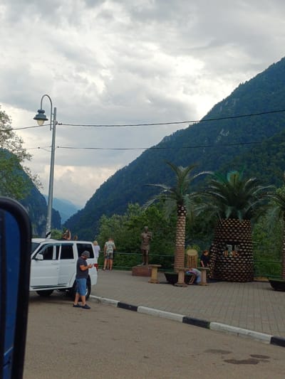 A group of people are gathered near a white vehicle on a paved area next to a road. The scene is set against a backdrop of a steep, tree-covered mountain under a cloudy sky. In the foreground, a man in a dark t-shirt and blue shorts stands beside the white vehicle, looking at his phone. Several other individuals are visible, some standing on the sidewalk and others near the vehicle. Behind them, a distinctive palm-tree-like structure with a patterned surface stands next to a paved walkway. One person is seated on a bench near the structure. The area appears to be a scenic overlook or tourist spot, with a lamppost also visible. The lighting suggests it is daytime, with overcast conditions. The overall impression is a casual stop during a trip, possibly in a mountainous or coastal region given the vegetation.