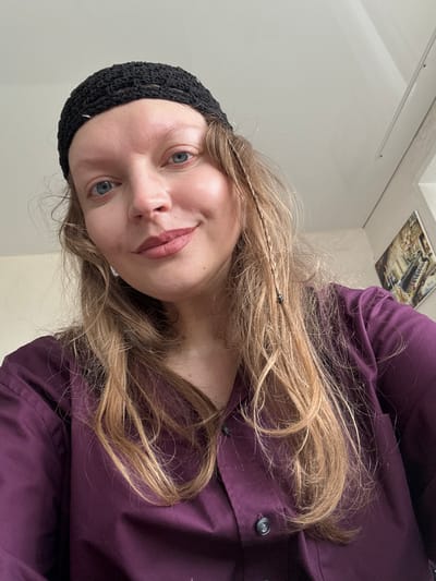 A close-up selfie taken indoors shows a person wearing a black crocheted beanie and a deep purple collared shirt. The person has light skin, blue eyes, and long, wavy blonde hair with a small braid. The background is a plain, light-colored wall with a framed picture on the right side, and a white ceiling. There is a window visible on the far right. The lighting appears to be natural and coming from the right. The scene does not contain any discernible elements that would indicate a specific location within the city of Postol, Russia.