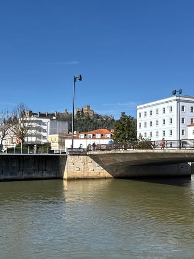 This image captures a daytime scene in Tomar, Portugal, viewed across the Nabão River. The foreground is dominated by the river, displaying a rippled, olive-green hue. A concrete and stone bridge spans the river, featuring a metal railing.

On the bridge, two individuals are visible on the right side walking left; one wears a bright orange top and dark trousers. Another individual is on the left side of the bridge, walking right. On the left riverbank, a paved promenade secured by a grey metal railing shows one pedestrian walking right.

Buildings line both banks. On the immediate right, a large, multi-story white building with numerous rectangular windows is prominent. On the left bank, a variety of structures include a white multi-story building with balconies, a yellow building, and several buildings topped with terracotta tile roofs.

Dominating the distant background, atop a heavily wooded hill, is the ancient Convento de Cristo and Castle of Tomar, characterized by its stone walls and towers. The sky above is clear and brilliant blue, with a faint, thin contrail visible. Two black streetlights, each equipped with multiple luminaire heads, are positioned in the mid-ground. Shadows are cast under the bridge, indicating a high sun position.
