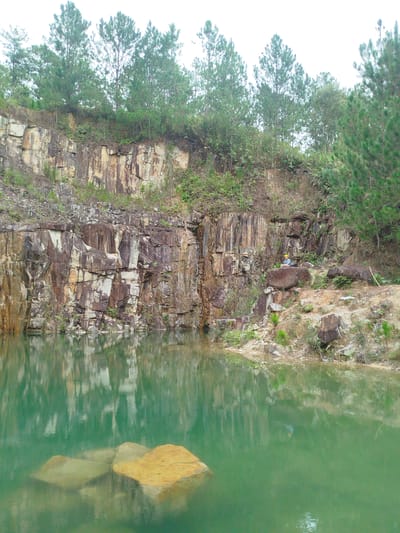 A quarry lake with sheer rock faces and a line of pine trees along the top edge is depicted. The water is a vibrant, reflective green, with submerged rocks visible near the foreground. A person, wearing a yellow hard hat and blue shirt, sits on a large rock on the right side of the frame, overlooking the lake. The rock face is a mosaic of earthy tones, with layers of sedimentary rock and patches of vegetation. The environment appears natural and somewhat remote, possibly in a rural area outside of Lán Tranh city, Vietnam.