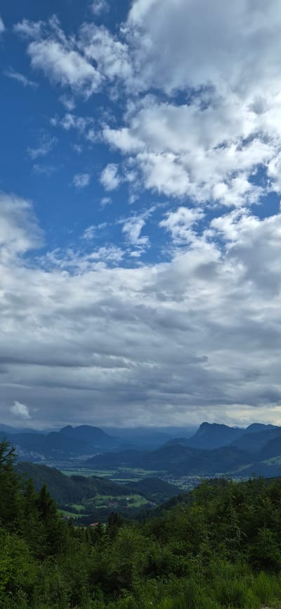 This vertical orientation photograph captures a vast mountainous landscape under a partially cloudy sky. The upper third of the image features a bright blue sky with scattered white cumulus clouds. Below this, a denser layer of grey and white clouds extends horizontally across the middle of the frame, partially obscuring the distant mountain peaks.
The mid-ground reveals a series of layered mountains, appearing in shades of deep blue and grey due to atmospheric haze. In the valley nestled between these mountains, an expanse of green fields and scattered residential buildings are visible, consistent with a rural settlement identified as being in or near Erl, Austria. Numerous small structures with red roofs are discernable within the valley. The foreground is dominated by a dense, vibrant green forest, composed of various trees and undergrowth, suggesting the viewpoint is from a elevated, forested area. No people or animals are visible. The lighting is diffused, indicating either late morning, afternoon, or an overcast day.