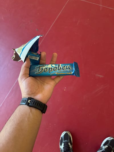 A person's hand is holding a partially unwrapped candy bar with a blue wrapper. The candy bar is labeled "Боровец" and "Тънък вафла". The background is a red, textured surface, possibly a floor or table. Two pairs of athletic shoes are visible at the bottom of the frame, one pair on the left and another on the right. The lighting suggests an indoor or shaded outdoor environment. There is no specific indication of the city of Dimovo, Bulgaria.