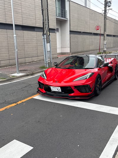 A bright red Chevrolet Corvette C8 is parked on a street in Nagoya, Japan. The car is angled towards the viewer, with its front end slightly to the left. The license plate is visible and reads "名古屋311 51". The car has a black roof and black accents, including a prominent rear spoiler. It is positioned on asphalt pavement marked with white pedestrian crossing lines and a yellow line. In the background, a modern building with horizontal wooden-like siding is visible. A utility pole stands to the right of the car, and a red and white fire alarm sign is mounted on it. The sky is overcast. The scene appears to be a typical urban street in Japan.