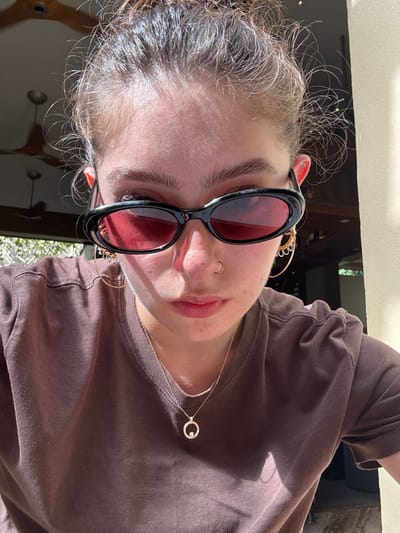 A woman wearing a brown t-shirt and black sunglasses with rose-tinted lenses poses in a close-up shot. She has fair skin, visible pores, and a silver necklace with a circular pendant. A small gold hoop earring peeks out near the sunglasses. Her hair is light brown and somewhat frizzy. The background features a door or window frame and outdoor foliage.