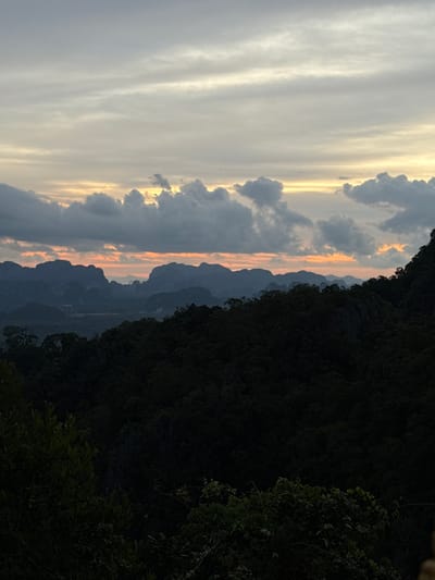 The image captures a panoramic view of a mountainous landscape at dusk. In the foreground, dense, dark green foliage of trees covers the lower portion of the frame, obscuring the ground. Beyond this, a series of silhouetted, jagged mountain peaks recede into the distance. The sky is a blend of soft grays and pale yellows, with streaks of orange and pink near the horizon, indicating the setting sun. Scattered, dark clouds drift across the sky, adding texture to the scene. The overall atmosphere is serene and natural, suggesting a remote or elevated vantage point, likely overlooking a valley or rural area within Ban Khao Tang, Thailand.