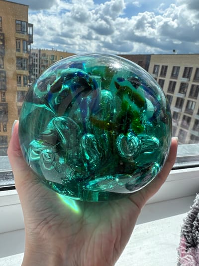 A hand is holding a large, transparent, green glass sphere filled with swirling dark blue and green pieces of glass. The sphere is positioned in front of a window, behind which modern apartment buildings and a cloudy sky are visible. The lighting suggests it is daytime.