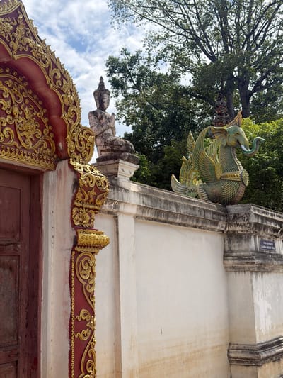 This media file captures an ornate architectural detail against a backdrop of lush trees and a cloudy sky. The primary subject is a decorative sculpture of a mythical creature, resembling a winged elephant with iridescent green and gold scales, perched on the edge of a weathered, stone-like structure. Its trunk curls upwards, and its wings are spread in a majestic pose. Below this sculpture, a richly decorated, deep red frieze with intricate gold ornamentation runs along the top of what appears to be a doorway. The goldwork features swirling patterns, floral motifs, and small circular embellishments.

The scene is outdoors, indicated by the natural light, the presence of a large tree with dense foliage dominating the upper left of the frame, and the visible sky with scattered clouds. The time of day appears to be daytime, with diffused light suggesting it might be overcast or early morning/late afternoon. The architecture suggests a cultural or religious site, likely a temple or traditional building, consistent with the provided location context of Ban Ko Klang, Thailand, known for its temples. The weathered stone and the somewhat aged appearance of the wooden door below the frieze add a sense of history and permanence to the scene. There are no visible people, and the focus is entirely on the static, decorative elements of the building and its natural surroundings. A small, rectangular sign with text is visible on the stone structure above the elephant sculpture, but the text is illegible at this resolution.