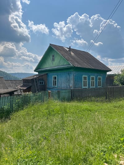 In Новомуллакаево, Russia, a traditional wooden house painted bright blue with green trim stands under a cloudy blue sky. The house has a dark, weathered roof and multiple windows with light-colored frames. A green wooden fence encloses the property, which is surrounded by tall, green grass and wildflowers. Adjacent to the main house, there is a smaller wooden structure with a sloped roof and a ladder leaning against it. In the background, a hilly, tree-covered landscape is visible, and power lines cross the sky above. The scene appears to be a rural residential setting.