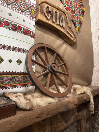 The image captures a rustic interior scene, likely an ethnographic display. A wooden wagon wheel leans against a fur pelt and a rough-hewn wooden post. A carved wooden sign is visible, partially obscured by a hand-woven textile with geometric patterns. Another colorful woven textile with fringes lies to the left of the sign. The scene is illuminated with natural light. Location unknown other than the context provided. The display appears to showcase traditional crafts and materials.