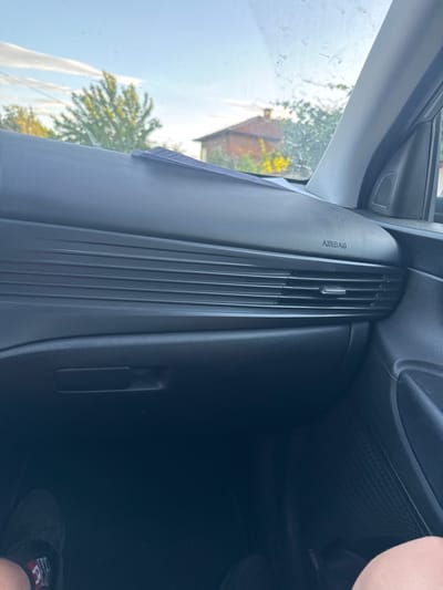 The image shows the interior of a car, specifically the dashboard area on the passenger side. "AIRBAG" is imprinted vertically on the dash. There are air vents in the dashboard. Outside the car window, a tree and the corner of a house or building are visible under a bright sky. The location is not discernible from the visual data provided. The city of Kaloyanovo, Bulgaria cannot be confirmed.