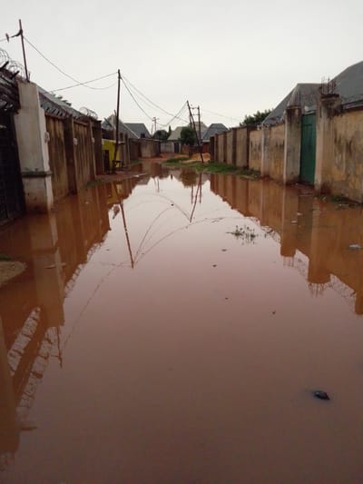 A flooded street in Rikina, Nigeria. The overcast sky suggests an early morning or late afternoon setting. The street is submerged in muddy, reddish-brown water, reflecting the surrounding buildings and utility poles. The buildings are primarily single-story structures with concrete walls and corrugated metal roofs. Barbed wire tops some of the walls, indicating security measures. Utility poles with tangled wires are visible along the street. A green door is set into one of the walls. There is a small patch of green grass on the left side of the street. No people are visible in the image. The scene depicts the aftermath of heavy rainfall.
