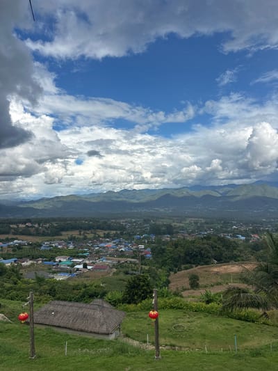 The image presents a panoramic landscape view of Ban Mai Saha Sampan, Thailand, captured from an elevated position under a dynamic sky. The upper approximately 60% of the frame features a bright blue sky partially covered by large, distinct cumulus clouds, varying in color from white to dense gray. A range of tree-covered mountains forms the horizon line in the background. Below the mountain range, the midground displays a valley containing the settlement of Ban Mai Saha Sampan, characterized by numerous residential structures with varied colored roofs, including blue, red, and gray, interspersed with green trees and cultivated fields. The foreground consists of a sloping, manicured grassy area. Centrally located in the lower foreground is a small, open-sided pavilion or hut with a distinctive multi-tiered, thatched or tiled roof. Two vertical wooden posts are present in the immediate foreground, positioned to the left and right of the hut, each supporting a spherical red lantern. A segment of a palm frond is visible on the far right edge of the frame. No individuals, actions, or specific interactions are observed within the scene.