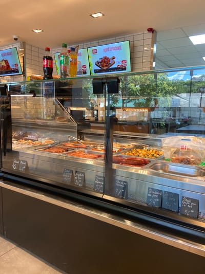 The image shows the interior of a food service establishment, possibly a deli or supermarket counter, in Soroca, Moldova. The scene features a refrigerated display case filled with various food items, including what appear to be sausages, meats, and possibly cooked dishes. Several price tags with handwritten text are visible in front of the food items. Above the display case, digital screens show advertisements for food items, one of which is labeled "POFTĂ DE VACANȚĂ" (Holiday Appetite). Bottles of Coca-Cola, Sprite, and another orange-colored beverage are positioned on a shelf above the display case, near the digital screens. The overall environment suggests a commercial setting with food for sale.