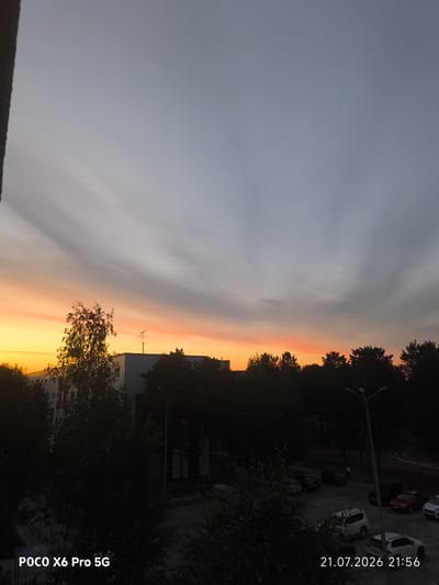 The image shows a dusk sky over the city of Kogalym, Russia. The sky displays a gradient of colors from orange and yellow at the horizon to grey at the top, with wispy clouds. Silhouetted against the sky are trees, a building with windows, and a television antenna. In the foreground, there are parked cars and a street lamp. The timestamp "21.07.2026 21:56" and the text "POCO X6 Pro 5G" are visible in the bottom corners of the image.