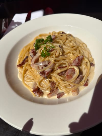 A top-down shot of a plate of pasta carbonara. The pasta is spaghetti, coated in a creamy sauce, and interspersed with bacon pieces. A small garnish of parsley sits on the left side of the plate. The pasta is served on a white plate. The photograph was taken outdoors, as a shadow is visible in the upper right corner of the plate. It is impossible to know if this image was taken in the city of Guildford, United Kingdom.
