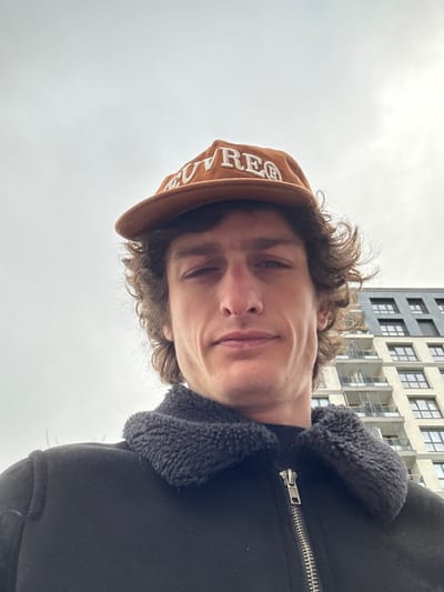 A male individual is depicted in a close-up, upward-angled selfie shot, positioned outdoors in Joinville-le-Pont, France. He appears to be in his late 20s or early 30s, with a slender face, prominent jawline, and curly brown hair visible beneath his headwear. His expression is neutral, with a slight upturn at the corners of his mouth, and he is looking directly at the camera.
He is wearing a brown baseball cap with the word "MUVREG" (or similar lettering) embroidered in cream-colored capital letters across the front. His attire includes a black jacket featuring a dark grey or black fleece/shearling collar and a visible silver-toned zipper pull on the front. A dark-colored garment, possibly a turtleneck, is visible beneath the jacket.
The background on the left consists of a uniformly overcast, light grey sky. To the right, a section of a multi-story modern building is visible, characterized by light-colored exterior panels, numerous dark-framed windows, and several balconies with transparent or semi-transparent railings. The architecture suggests a contemporary residential or office structure. No other specific ground-level features or landmarks of Joinville-le-Pont are discernible.