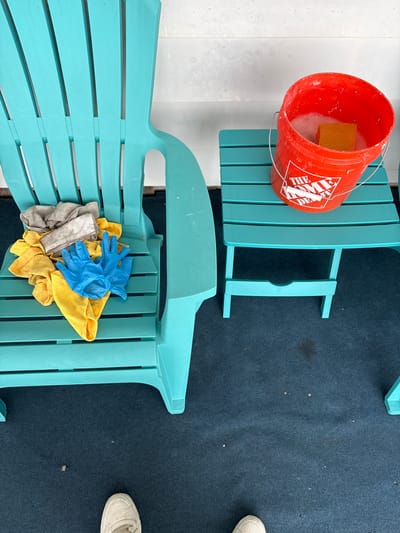 This media file captures an overhead view of an outdoor cleaning setup, likely on a patio or deck in Hampton, USA.
The main subjects are two teal-colored Adirondack chairs. The top chair has a bright orange Home Depot bucket resting on its side, filled with soapy water and a yellow sponge. The bucket prominently displays "THE HOME DEPOT" logo. The bottom chair holds a pile of cleaning supplies, including a pair of blue rubber gloves, several yellow microfiber cloths, and a gray cleaning rag.
The scene suggests an activity of cleaning the outdoor furniture. The presence of the bucket with soapy water, sponge, gloves, and cloths all indicate that the chairs are either being cleaned or have just been cleaned.
Notable details include the ground surfaces: a light-colored, possibly white, tiled or paved area on the left, and a dark blue outdoor carpet or mat covering the right side. A person's foot, wearing a white sneaker, is partially visible on the carpet in the bottom right corner of the frame. The lighting appears bright and even, characteristic of daytime.
