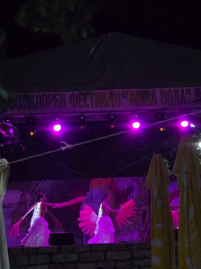 The image captures a nighttime outdoor performance at what appears to be a cultural festival. The scene is dominated by vibrant purple and pink stage lighting that illuminates the performers and a vertical banner.

The main subjects are two performers dressed in elaborate costumes. One performer, in the foreground and to the right, is dressed in a white, flowing costume that is lit with small lights. They are positioned dynamically, with one arm extended and the other holding a pink fan. This performer is likely in mid-dance or acrobatic pose. Another performer, partially visible in the background and to the right, also seems to be in motion, possibly holding fans or props that are highlighted by the lighting.

Objects visible include large, yellow umbrellas or canopies stretched out overhead, providing a backdrop and perhaps shelter. There are also lights mounted on a vertical pole or structure, casting beams of light. A brick wall is visible on the far right, suggesting an outdoor setting with some permanent structures.

The activity taking place is clearly a performance, likely a dance or artistic display, given the costumes and dynamic poses of the performers. The lighting suggests a theatrical or festive atmosphere.

Notable details include the nighttime setting, indicated by the dark background and artificial lighting. The intense purple and pink lights create a dramatic and celebratory mood. The text on the vertical banner is in Cyrillic script and appears to read "ФОЛКЛОРЕН ФЕСТИВАЛ 'ЖИВА ВОДА'" which translates to "FOLKLORE FESTIVAL 'LIVING WATER'". This confirms the event is a folklore festival. The location context provided is Gara Oreshets, Bulgaria, which aligns with the Cyrillic text. The overall impression is one of lively entertainment and cultural celebration.