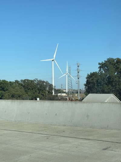 The image shows a clear blue sky over a rural landscape. In the middle ground, two large white wind turbines are visible, with their blades extended. To the right of the turbines, there is a tall electrical transmission tower with multiple wires. A line of dense green trees runs horizontally across the middle of the frame, partially obscuring some of the structures behind them. In the foreground, a weathered concrete barrier runs along the bottom of the image. A metal-roofed shed or building is visible behind the barrier and to the right of the trees. The scene appears to be an open area, possibly on the outskirts of Fairfield, California, given the presence of wind turbines and transmission infrastructure.